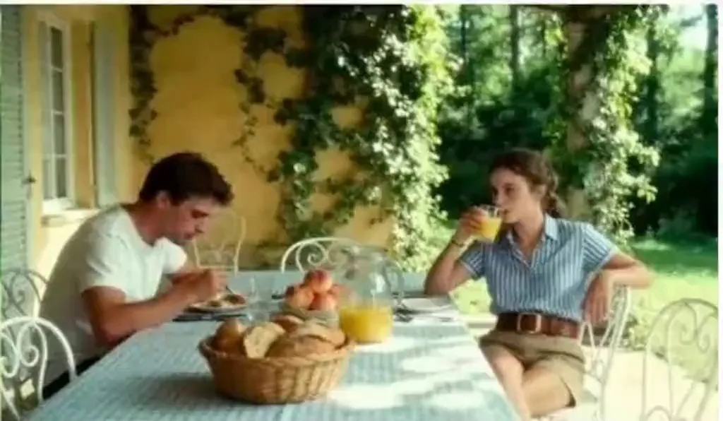 kling 3.0 audio sync | European villa outdoor terrace scene. At a table with blue and white checkered tablecloth, a young white woman in a blue and white striped short-sleeve shirt and khaki shorts with brown belt sits barefoot. Across from her is a young white man in a white T-shirt. Camera pushes in as the woman swirls juice in her glass, gazing at the distant trees, saying 'These trees will turn yellow in a month, won't they?' Close-up of the man looking down, saying 'but they'll be green again next summer.' The woman turns her head, smiling at the man across from her, saying 'Are you always this optimistic? Or just about summer?' The man looks up at her and says 'Only about summers with you.'